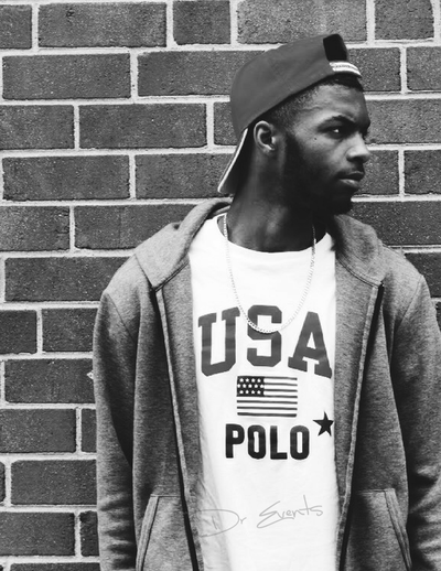 Black and white photograph of a young man with a USA t-shirt on standing against a brick wall.